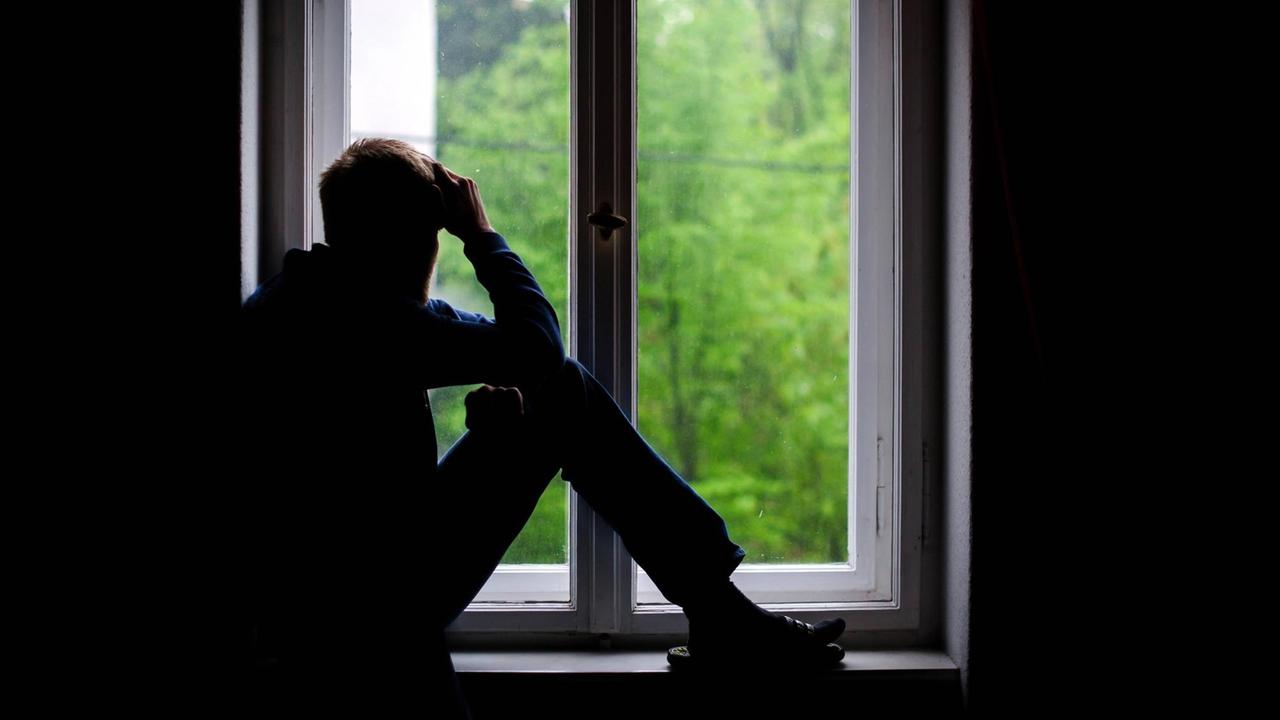 Ein Mann sitzt in einem dunklen Raum an einem Fenster und schaut hinaus Ein Mann sitzt in einem dunklen Raum an einem Fenster und schaut hinaus