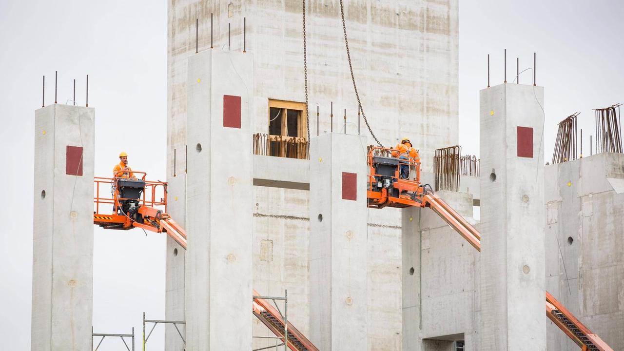 Die Bosch-Baustelle in Dresden. Zwischen grauen Betonpfeilern stehen Bauarbeiter auf orange farbenen Hebebühnen Die Bosch-Baustelle in Dresden. Zwischen grauen Betonpfeilern stehen Bauarbeiter auf orange farbenen Hebebühnen
