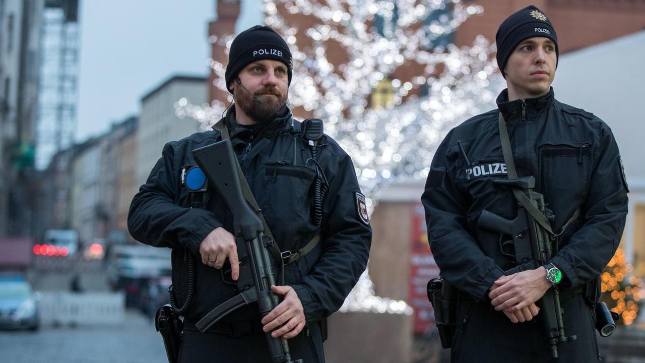 Zwei Polizisten in schwarzen Uniformen und mit Maschinengewehren stehen vor einem mit Lichterketten geschmückten Baum vor dem Eingang des Weihnachtsmarktes.