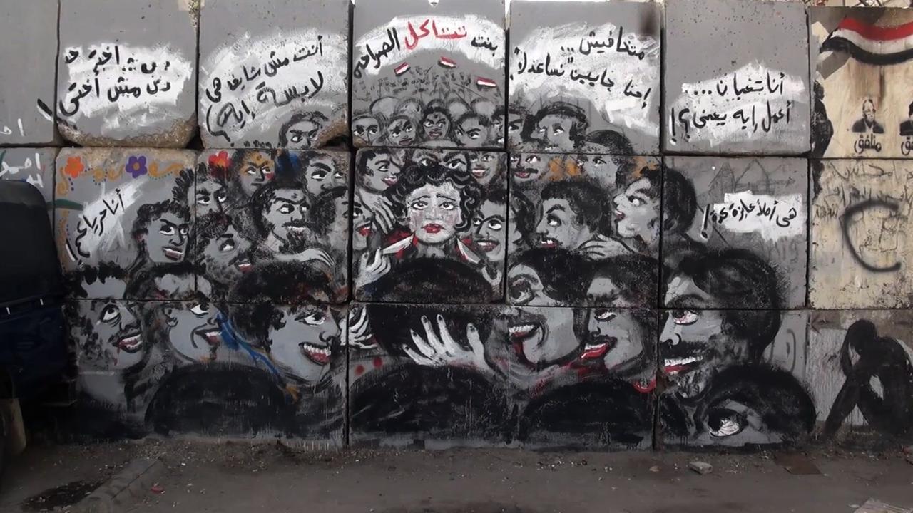 Das Still aus "As I Want" zeigt ein Graffiti mit vielen Männern, die eine Frau im Zentrum bedrohen.