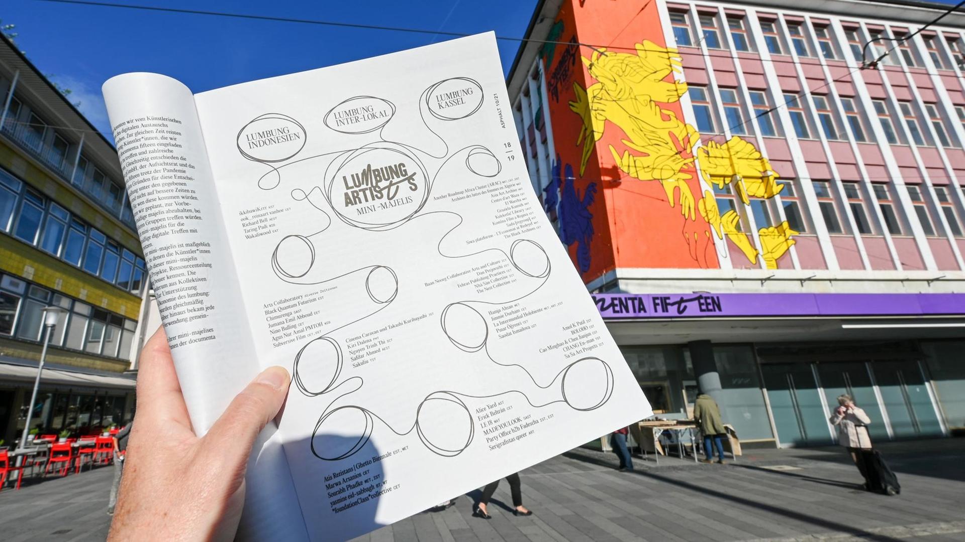 Eine Hand hält die Straßenzeitung "Asphalt", aufgeschlagen auf der Seite mit der kompletten Liste der ausstellenden Künstlerinnen und Künstler der Documenta 15, vor dem "ruruHaus".