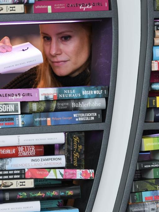 Die Frau stapelt Bücher in zwei Fenstern; ihr Kopf ist durch ein letztes verbliebenes Loch zwischen den Büchern zu sehen.