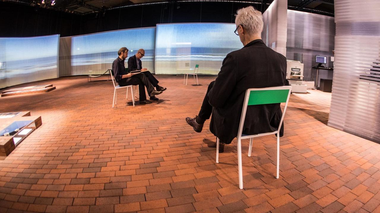 Drei Menschen sitzen auf Stühlen auf den Pflastersteinen und lesen; im Hintergrund ist Meeresbrandung auf Vorhänge projeziert. Drei Menschen sitzen auf Stühlen auf den Pflastersteinen und lesen; im Hintergrund ist Meeresbrandung auf Vorhänge projeziert.
