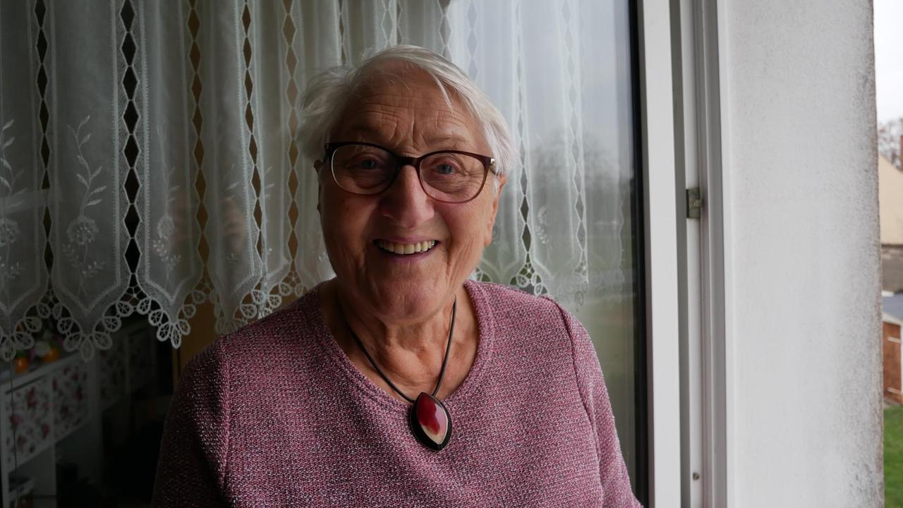 Eine ältere Frau im lilafarbenen Pullover und dunkel umrandeter Brille sitzt auf einem Balkon.