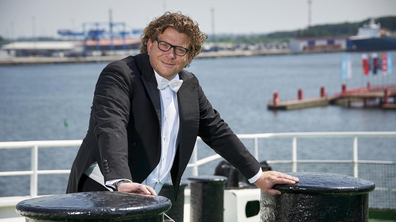 Ein Mann im Frack steht auf einem Schiff im Hafen und stützt sich mit seinen Händen jeweils auf einen Metallsockel, der für Leinen bestimmt. Ein Mann im Frack steht auf einem Schiff im Hafen und stützt sich mit seinen Händen jeweils auf einen Metallsockel, der für Leinen bestimmt.