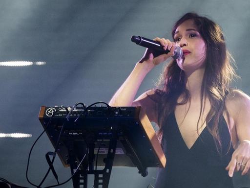 Eine junge Frau mit langen braunen Haaren steht mit Mikrofon, das sie mit der rechten Hand vor ihren Mund hält, auf einer Bühne im Scheinwerferlicht.