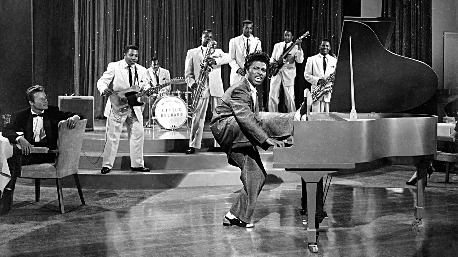 Eine historische Aufnahme in scharz-weiß zeigt den Musiker Little Richard am Klavier bei einer Fernsehaufzeichnung. Er hebt sein rechtes Bein. Im Hintergrund ist seine sechsköpfige Band zu sehen.