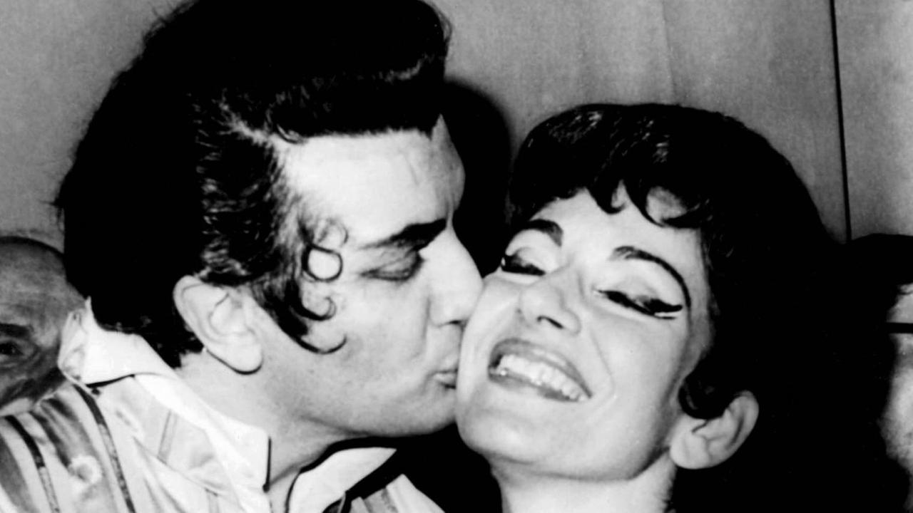 Schwarz-weiß Foto. Der Tenor Franco Corelli gibt der Sopranistin Maria Callas, beide sind stark geschminkt und tragen Bühnenkostüme, einen Kuß auf die rechte Wange