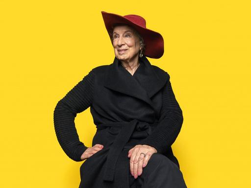 Ein Porträtfoto zeigt Margaret Atwood mit rotem Hut vor einem gelben Hintergrund. Geboren 1939 in Ottawa, gehört zu den bedeutendsten Erzählerinnen unserer Zeit.