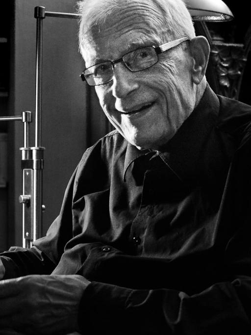 Hermann Schmitz, ein alter Herr mit weißem Haar und Brille, sitzt an seinem Schreibtisch und lächelt in die Kamera.