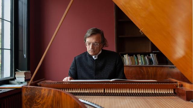 Ein älterer Mann mit grauen langen Haaren sitzt vertieft in sein Spiel an einem historischen Klavier.