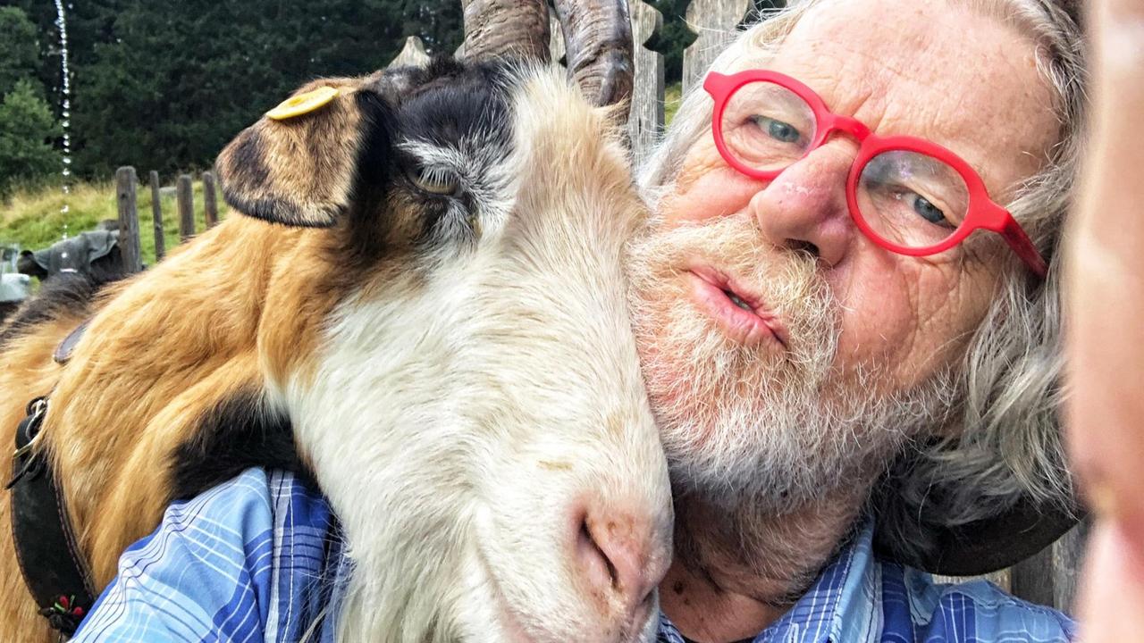 Eine Ziege legt ihren Kopf auf die Schulter eines älteren, grauhaarigen Mannes mit Vollbart und roter Brille.