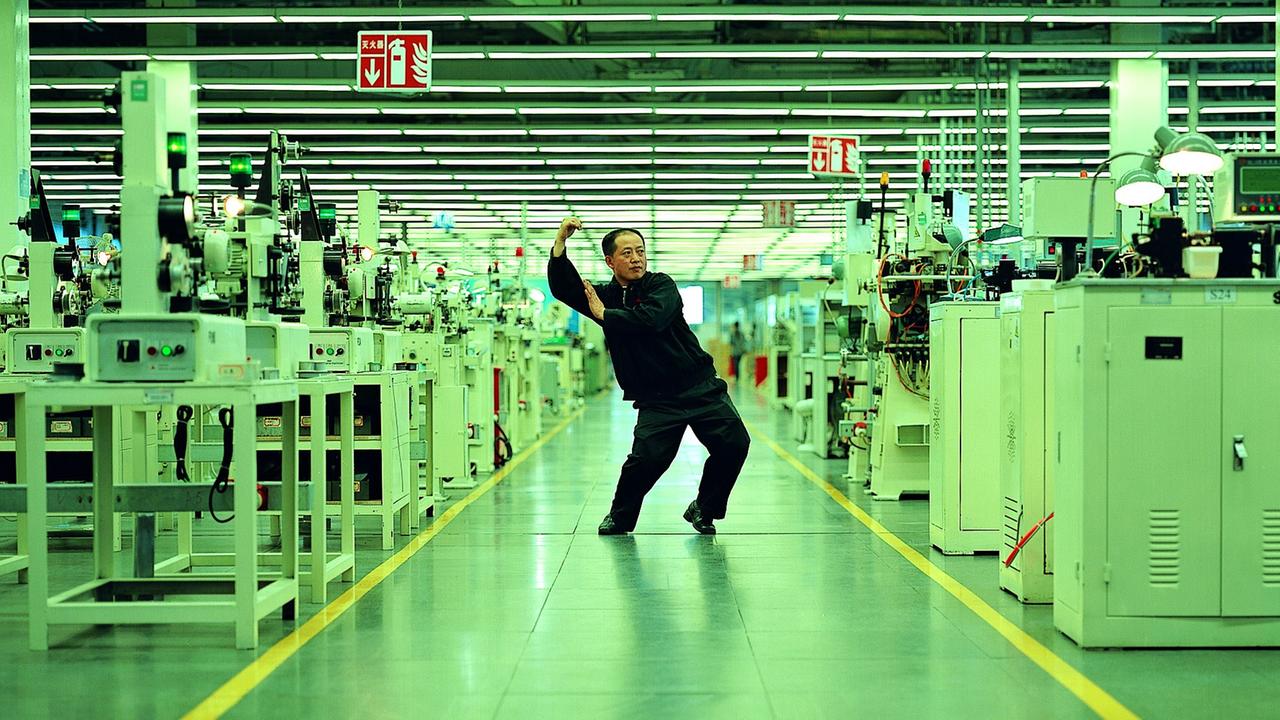 Ein asiatischer Mann in einem dunklen Overall steht inmitten von Maschinen. Cao Fei, "Whose Utopia", 2006.