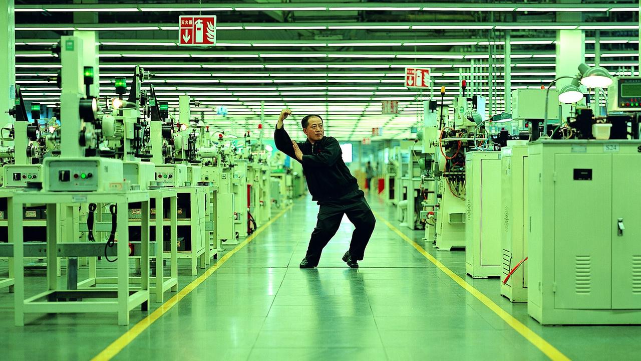 Ein asiatischer Mann in einem dunklen Overall steht inmitten von Maschinen. Cao Fei, "Whose Utopia", 2006. Ein asiatischer Mann in einem dunklen Overall steht inmitten von Maschinen. Cao Fei, "Whose Utopia", 2006.