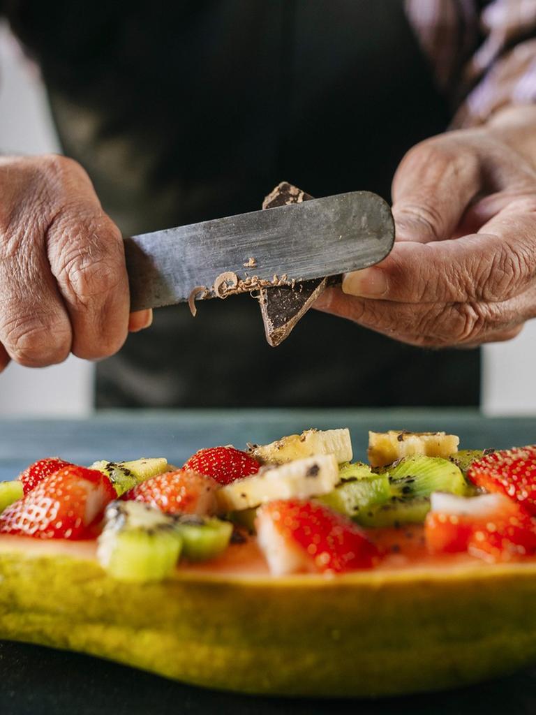Nahaufnahme von den Händen eines alten Mannes, der Schokolade über eine Papayahälfte raspelt.