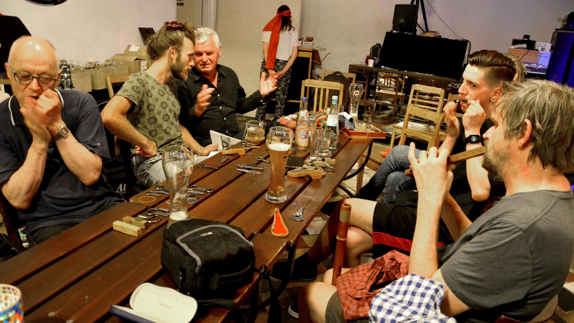 An einem Kneipentisch sitzen einige Menschen und unterhalten sich angeregt. Ein Herr im Vordergrund spielt auf einer Maultrommel. An einem Kneipentisch sitzen einige Menschen und unterhalten sich angeregt. Ein Herr im Vordergrund spielt auf einer Maultrommel.
