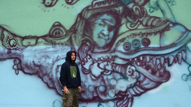 Der Street-Art-Künstler WON ABC steht in einem Kapuzenpulli, Armyhose und schweren Arbeiterboots vor einer von ihm besprühten Museumswand.