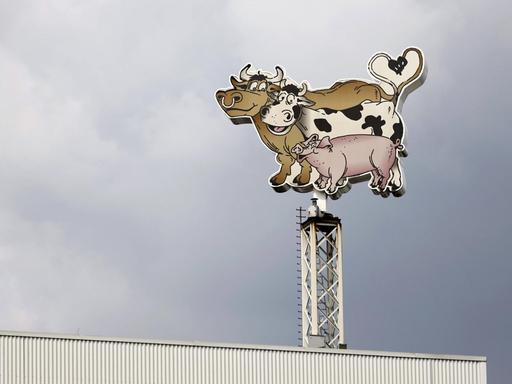 Logo der Lebensmittelfirma Tönnies bestehend aus einem Bullen, einer Kuh und einem Schwein am Hauptsitz in Rheda-Wiedenbrück vor dunklen Wolken.
