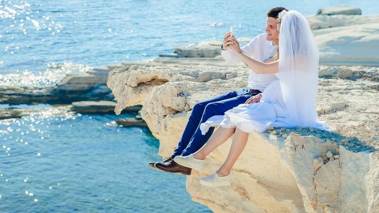 Ein Hochzeitspaar sitzt auf einem Felsen am Strand und macht ein Selfie Ein Hochzeitspaar sitzt auf einem Felsen am Strand und macht ein Selfie