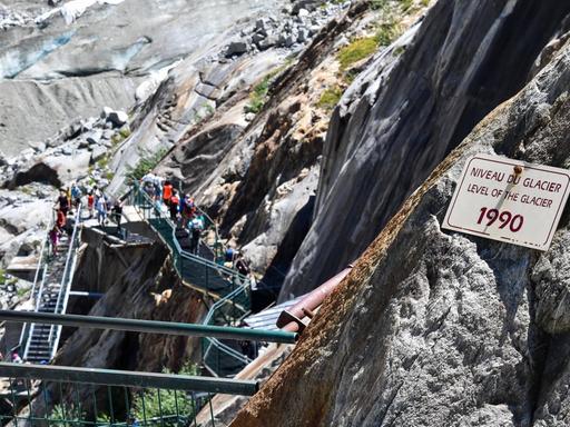 Eine Eisentreppe führt an kahlen, gerölligen Felswänden hinunter. An einem Felsen ist ein Schild angebracht, das darauf hinweist, dass 1990 hier die Decke des Gletschers war.