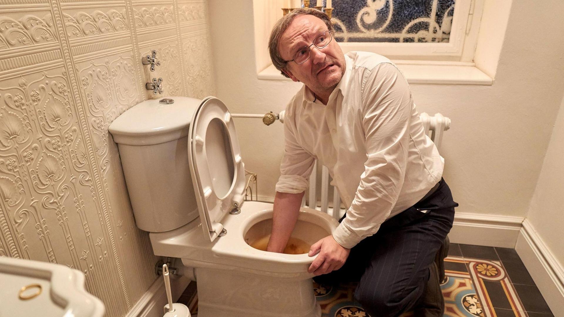 Szenefoto aus dem Fernsehfilm. Josef Asch, gespiel von Rainer Bock greift in einem Badezimmer mit der rechten Hand in die Toilettenschüssel. Szenefoto aus dem Fernsehfilm. Josef Asch, gespiel von Rainer Bock greift in einem Badezimmer mit der rechten Hand in die Toilettenschüssel.