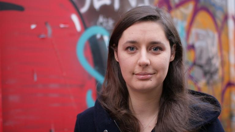 Katharina Nocun, Bürgerrechtlerin und Netzaktivistin. Eine junge Frau mit langen Haaren steht vor einer mit Graffiti besprühten Wand. 