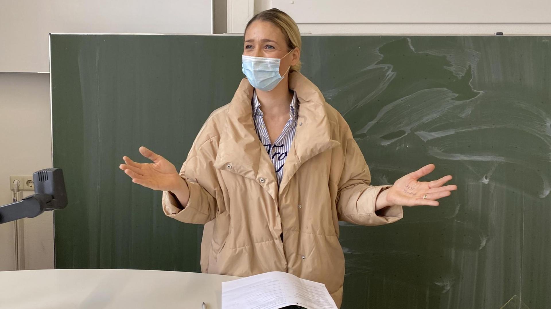 Eine Lehrerin mit Winterjacke und Mundschutz steht vor der Tafel und gestikuliert Eine Lehrerin mit Winterjacke und Mundschutz steht vor der Tafel und gestikuliert