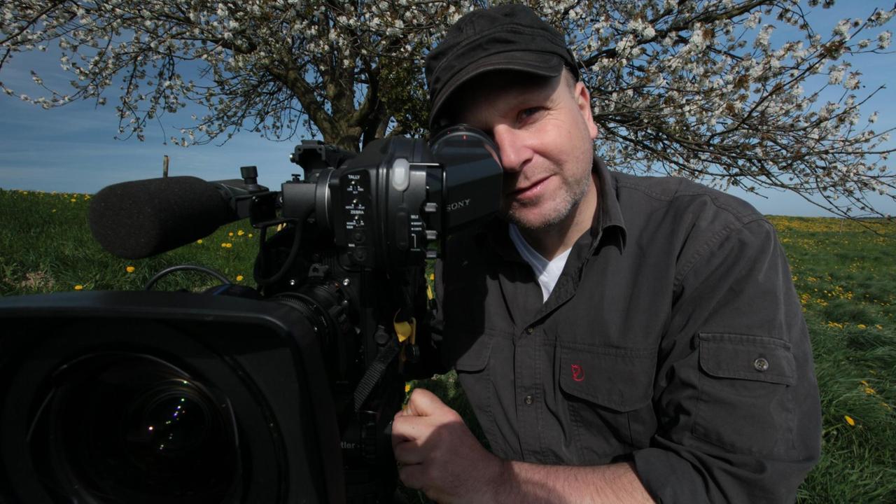 Der Tierfilmer Christoph Hauschild hinter seiner Kamera auf einer grünen Wiese mit einem Baum im Hintergrund Der Tierfilmer Christoph Hauschild hinter seiner Kamera auf einer grünen Wiese mit einem Baum im Hintergrund