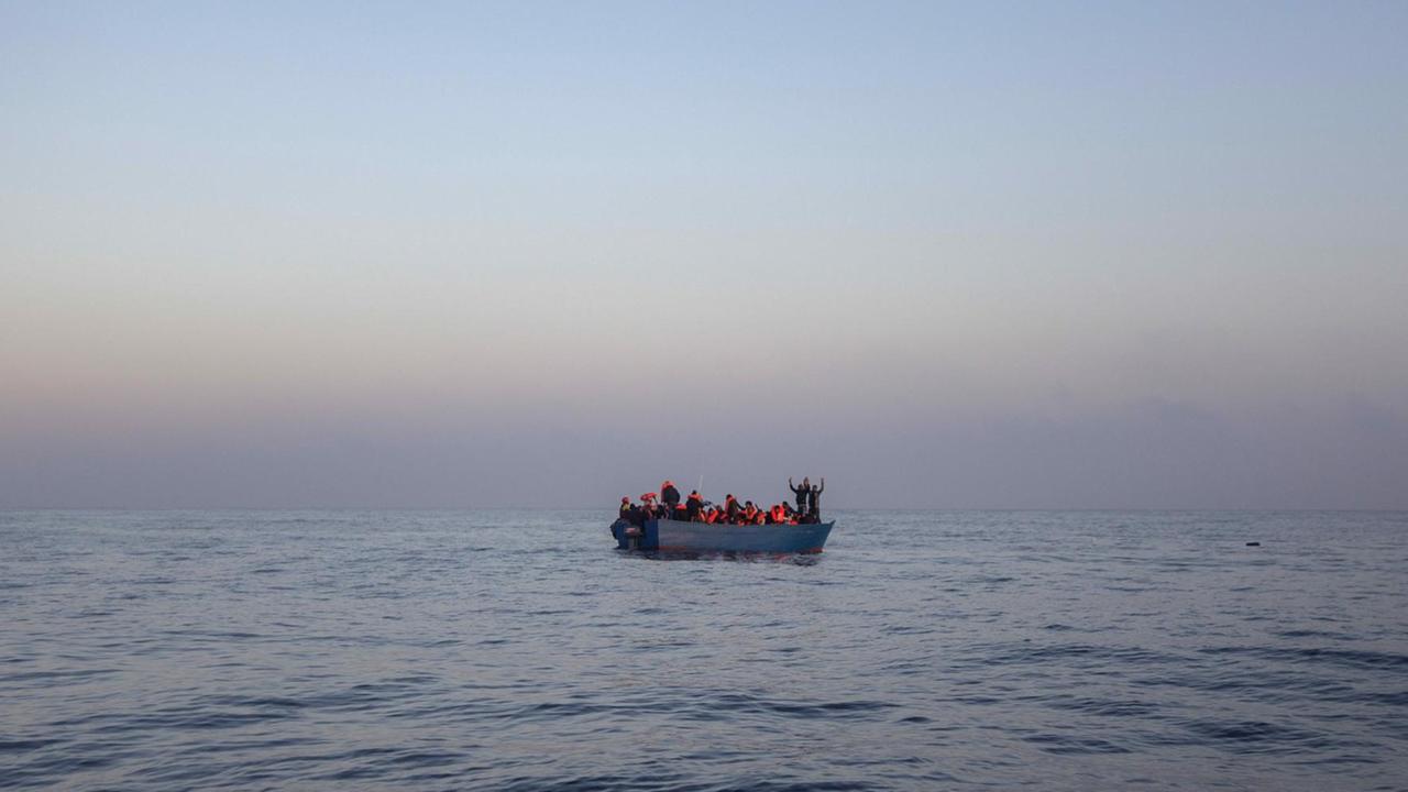 Bootsflüchtlinge aus Afrika und Asien winken auf einem Holzboot im Mittelmeer, 14 Meilen nördlich von Sabratha, Libyen, während sie auf ihre Rettung durch eine NGO warten Bootsflüchtlinge aus Afrika und Asien winken auf einem Holzboot im Mittelmeer, 14 Meilen nördlich von Sabratha, Libyen, während sie auf ihre Rettung durch eine NGO warten