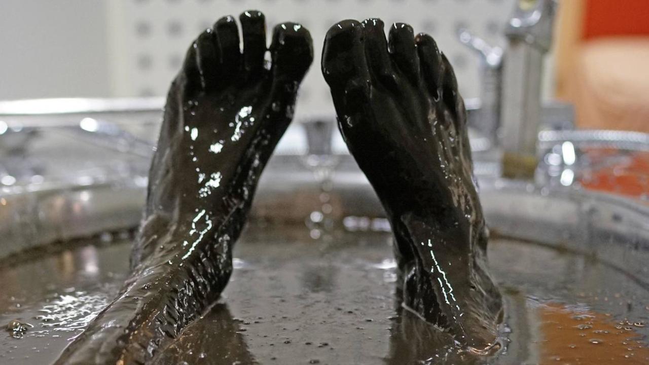 Zwei Füße, auf die der Betrachter schaut, als wären es seine eigenen, während er in der Badewanne liegt. Das Bad ist allerdings ein Moorbad, so dass die Füße ganz braun sind.