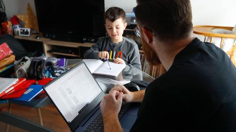 Ein Mann sitzt mit einem Kind zu Hause. Das Kind mach Schulaufgaben, der Mann sitzt am Rechner.
