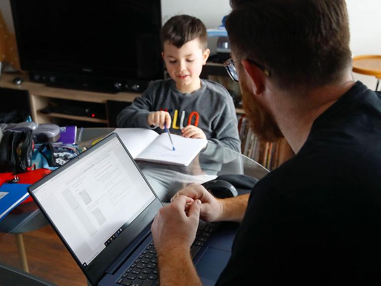 Ein Mann sitzt mit einem Kind zu Hause. Das Kind mach Schulaufgaben, der Mann sitzt am Rechner.