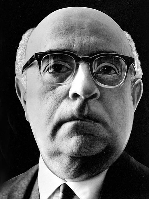 Ein Nahaufnahme von Theodor W. Adorno vor schwarzem Hintergrund. Er trägt eine Hornbrille und schaut aufmerksam dem Betrachter entgegen.