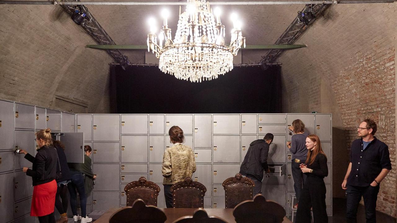 Die Aufnahme zeigt Theatergäste bei der Performance von Tania El Khoury. Sie schließen Türen in einer raumgroßen Tresorwand auf, zwei Theatergäste stecken die Köpfe in die Schließfächer. Die Szenerie wird von einem prächtigen Kronleuchter in der Mitte des Raumes beleuchtet. Die Aufnahme zeigt Theatergäste bei der Performance von Tania El Khoury. Sie schließen Türen in einer raumgroßen Tresorwand auf, zwei Theatergäste stecken die Köpfe in die Schließfächer. Die Szenerie wird von einem prächtigen Kronleuchter in der Mitte des Raumes beleuchtet.