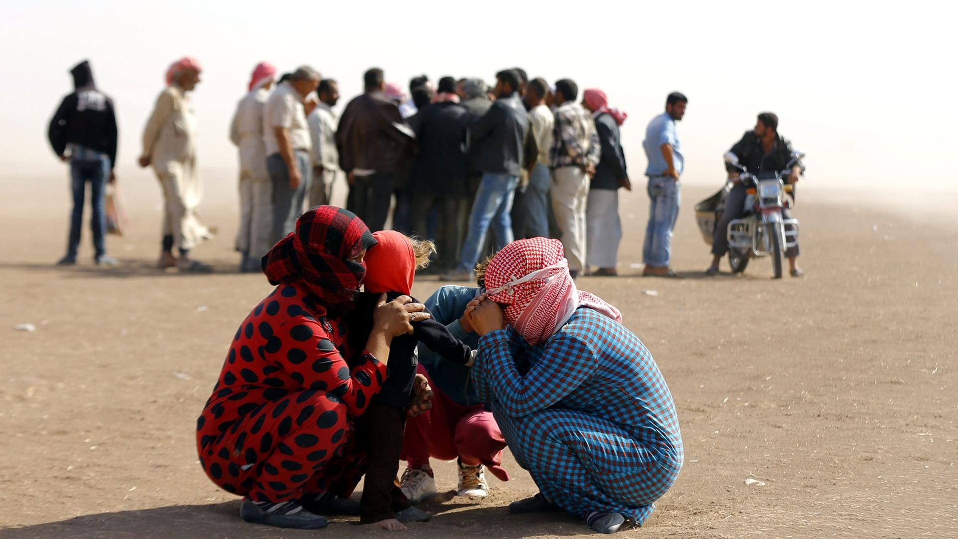 Syrische Flüchtlinge an der Grenze nahe der türkischen Stadt Sanliurfa: Zwei Frauen und zwei Kinder kauern zusammen und schützen sich mit Kopftüchern gegen den Wind. Dahinter steht eine Gruppe von Flüchtlingen Schlange. 