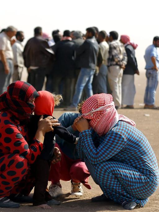 Syrische Flüchtlinge an der Grenze nahe der türkischen Stadt Sanliurfa: Zwei Frauen und zwei Kinder kauern zusammen und schützen sich mit Kopftüchern gegen den Wind. Dahinter steht eine Gruppe von Flüchtlingen Schlange. 