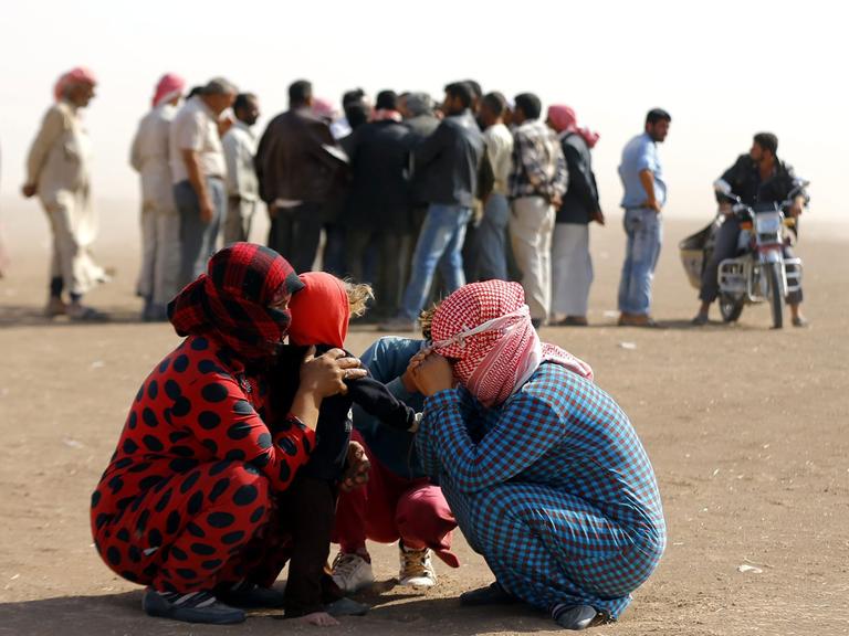 Syrische Flüchtlinge an der Grenze nahe der türkischen Stadt Sanliurfa: Zwei Frauen und zwei Kinder kauern zusammen und schützen sich mit Kopftüchern gegen den Wind. Dahinter steht eine Gruppe von Flüchtlingen Schlange. 
