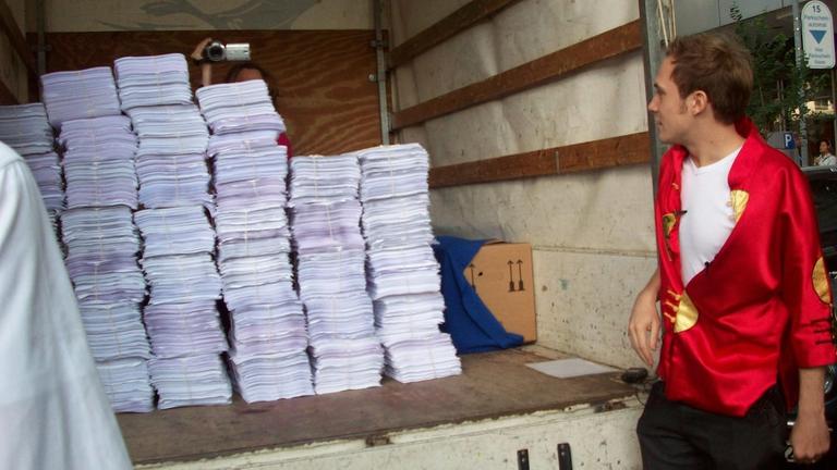 Der Komponist und Aktionskünstler Johannes Kreidler steht neben der Ladefläche eines Transporters mit Stapeln von Papier Der Komponist und Aktionskünstler Johannes Kreidler steht neben der Ladefläche eines Transporters mit Stapeln von Papier