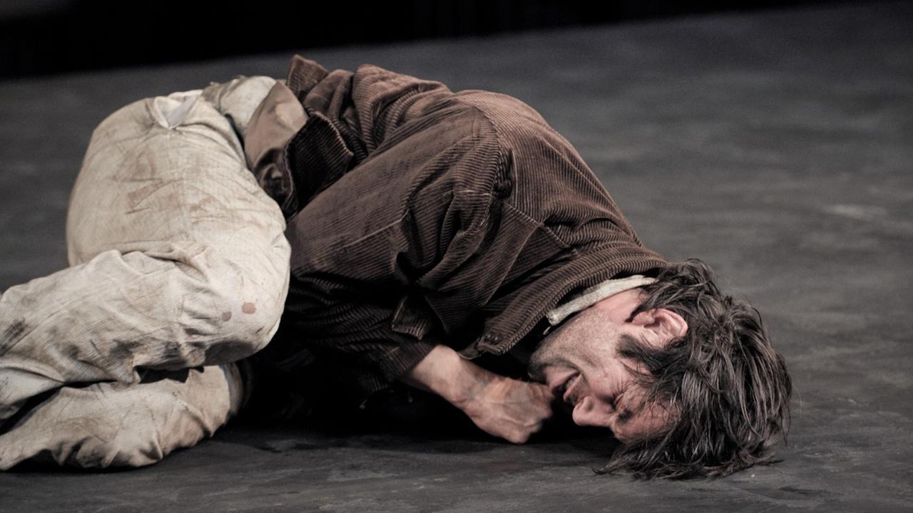 Bühnenszene, in der ein Mann (Max Simonischek als Georg) zusammengekauert auf dem Boden liegt Bühnenszene, in der ein Mann (Max Simonischek als Georg) zusammengekauert auf dem Boden liegt