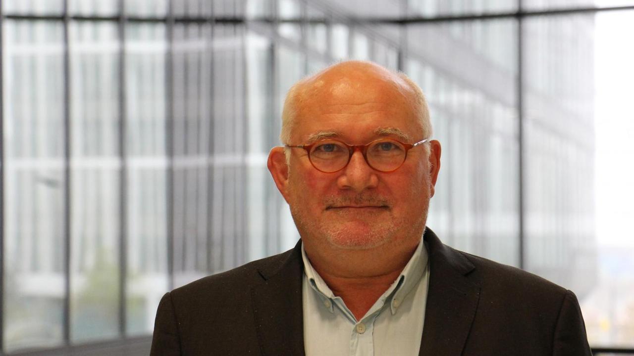 Thomas Krüger blickt in Richtung der Kamera. Er träge eine dunkle Jacke und ein helles Hemd, der Blick durch die Fenster hinter ihm zeigt einen weiteren Flügel des Gebäudes, in dem Krüger steht. Thomas Krüger blickt in Richtung der Kamera. Er träge eine dunkle Jacke und ein helles Hemd, der Blick durch die Fenster hinter ihm zeigt einen weiteren Flügel des Gebäudes, in dem Krüger steht.