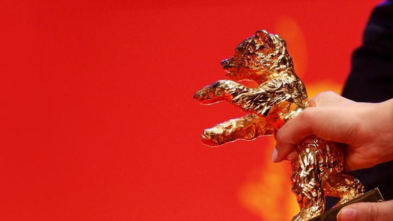 Detail einer weblichen Hand vor rotem Hintergrund, die einen Goldenen Bären festhält. Detail einer weblichen Hand vor rotem Hintergrund, die einen Goldenen Bären festhält.