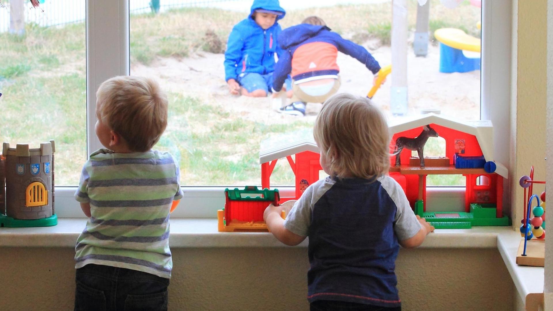 Zwei Kinder stehen am Fenster einer Kita und gucken anderen Kindern draußen beim Spielen zun Zwei Kinder stehen am Fenster einer Kita und gucken anderen Kindern draußen beim Spielen zun