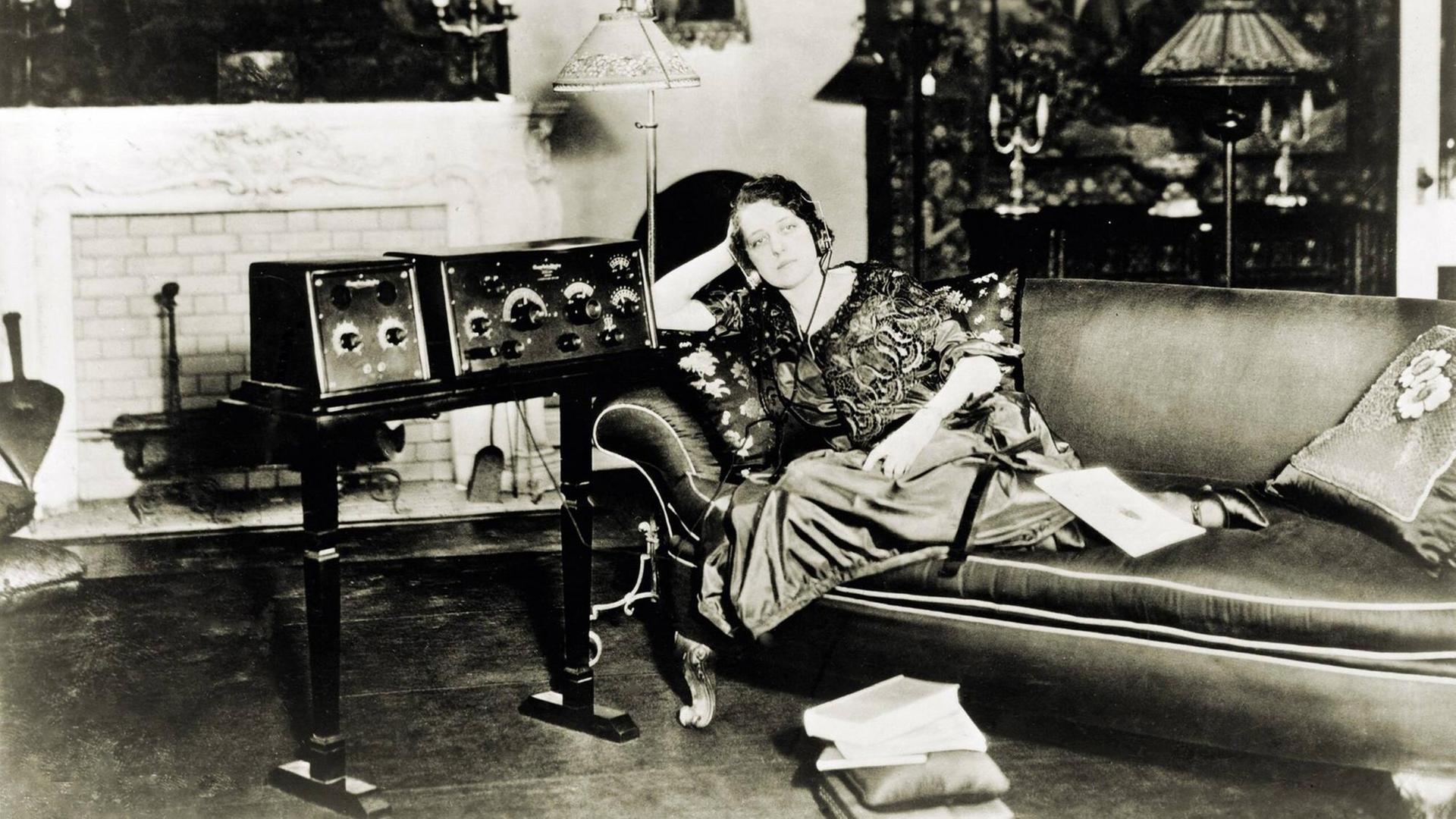 Eine Frau liegt 1923 auf ihrem Sofa und hört versunken Radio über ihre Kopfhörer, die an ein großes Rundfunkgerät angeschlossen sind.