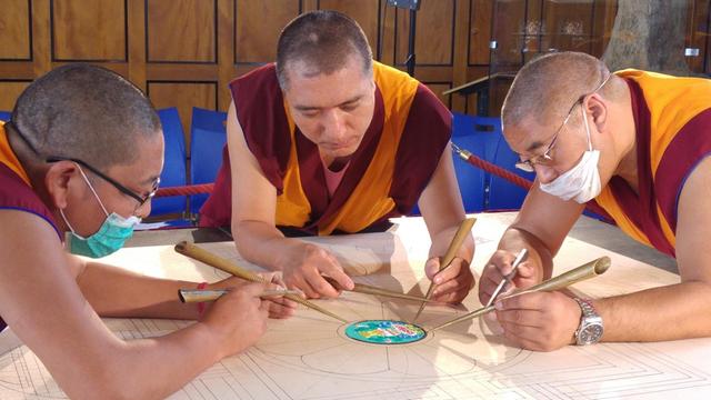 Mönche beim Erstellen eines Sandmandalas. Dabei tragen sie bunten Sand mit speziellen Metalltüllen auf die Holzplatte mit dem Mandala auf.