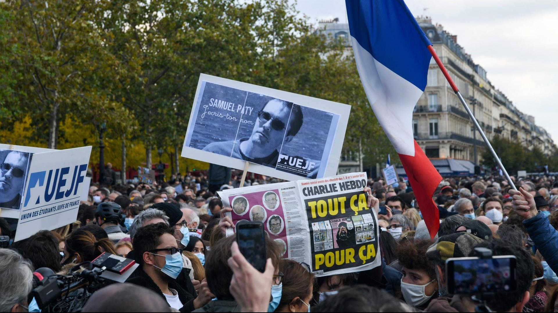 In Frankreich demonstrieren viele Menschen in Erinnerung an den ermordeten Lehrer Samuel Paty. Sie zeigen ein Bild von ihm, eine Frankreich-Fahne und eine Zeitung, auf der eine Mohammed-Karikatur zu erkennen ist. In Frankreich demonstrieren viele Menschen in Erinnerung an den ermordeten Lehrer Samuel Paty. Sie zeigen ein Bild von ihm, eine Frankreich-Fahne und eine Zeitung, auf der eine Mohammed-Karikatur zu erkennen ist.