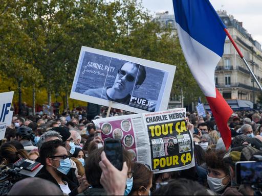 In Frankreich demonstrieren viele Menschen in Erinnerung an den ermordeten Lehrer Samuel Paty. Sie zeigen ein Bild von ihm, eine Frankreich-Fahne und eine Zeitung, auf der eine Mohammed-Karikatur zu erkennen ist.