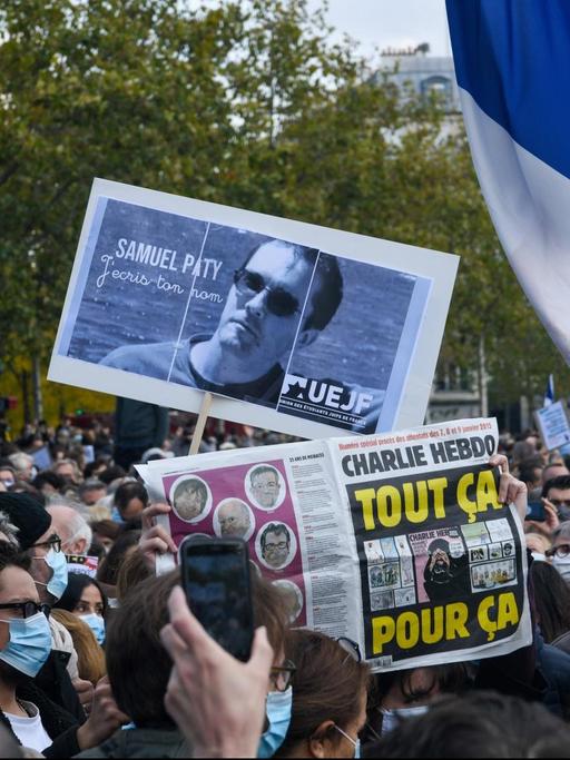 In Frankreich demonstrieren viele Menschen in Erinnerung an den ermordeten Lehrer Samuel Paty. Sie zeigen ein Bild von ihm, eine Frankreich-Fahne und eine Zeitung, auf der eine Mohammed-Karikatur zu erkennen ist.