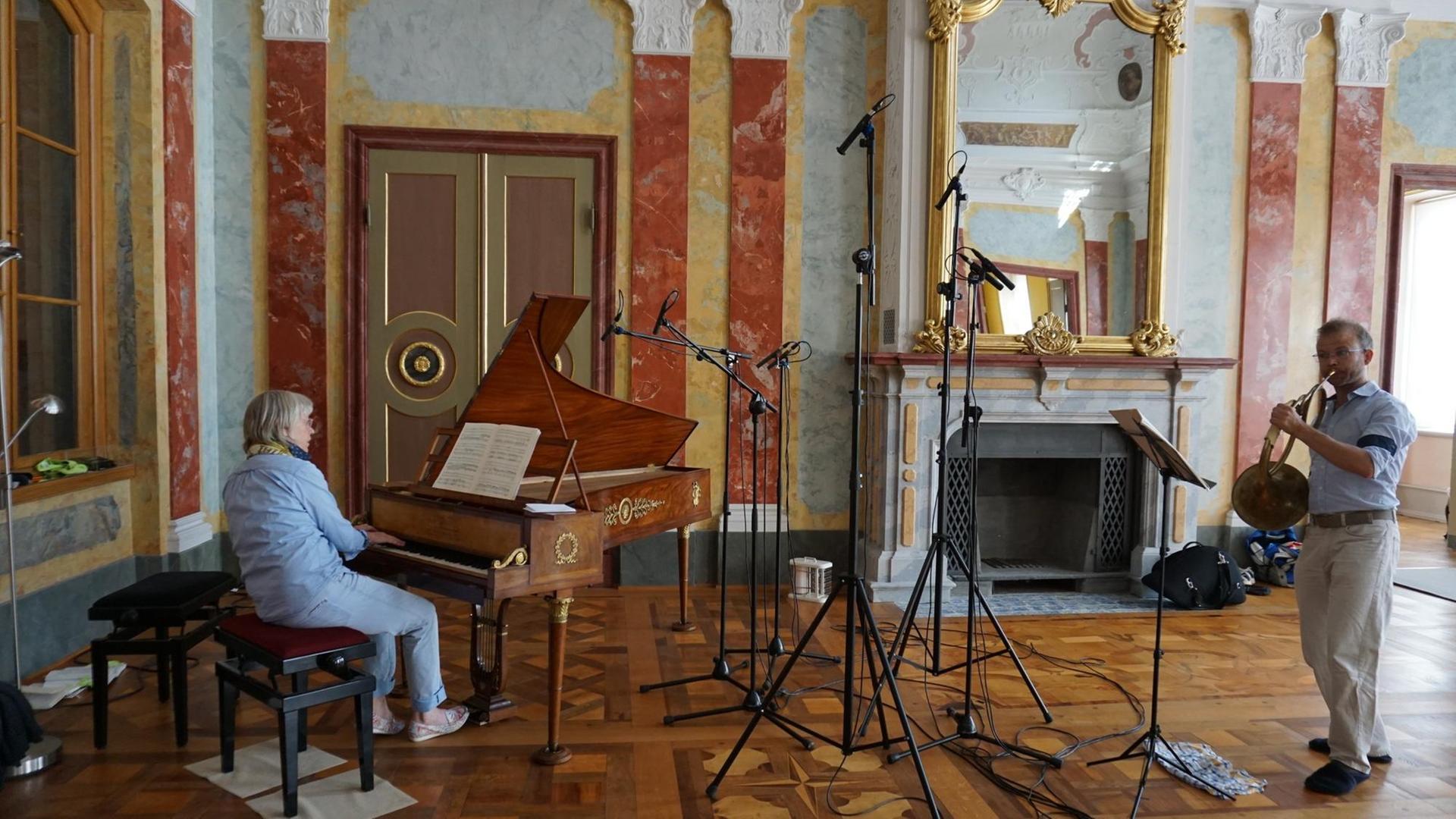 In einem barocken Festsaal spielen eine Musikerin an einem historischen Hammerklavier und ein Hornist zwischen Mikrofonstativen.