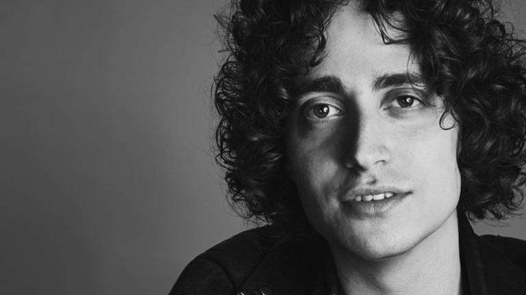 Ein junger Mann mit wilden dunklen Locken in Lederjacke schaut direkt mit leicht geöffnetem und lächelndem Mund in die Kamera. Ein junger Mann mit wilden dunklen Locken in Lederjacke schaut direkt mit leicht geöffnetem und lächelndem Mund in die Kamera.
