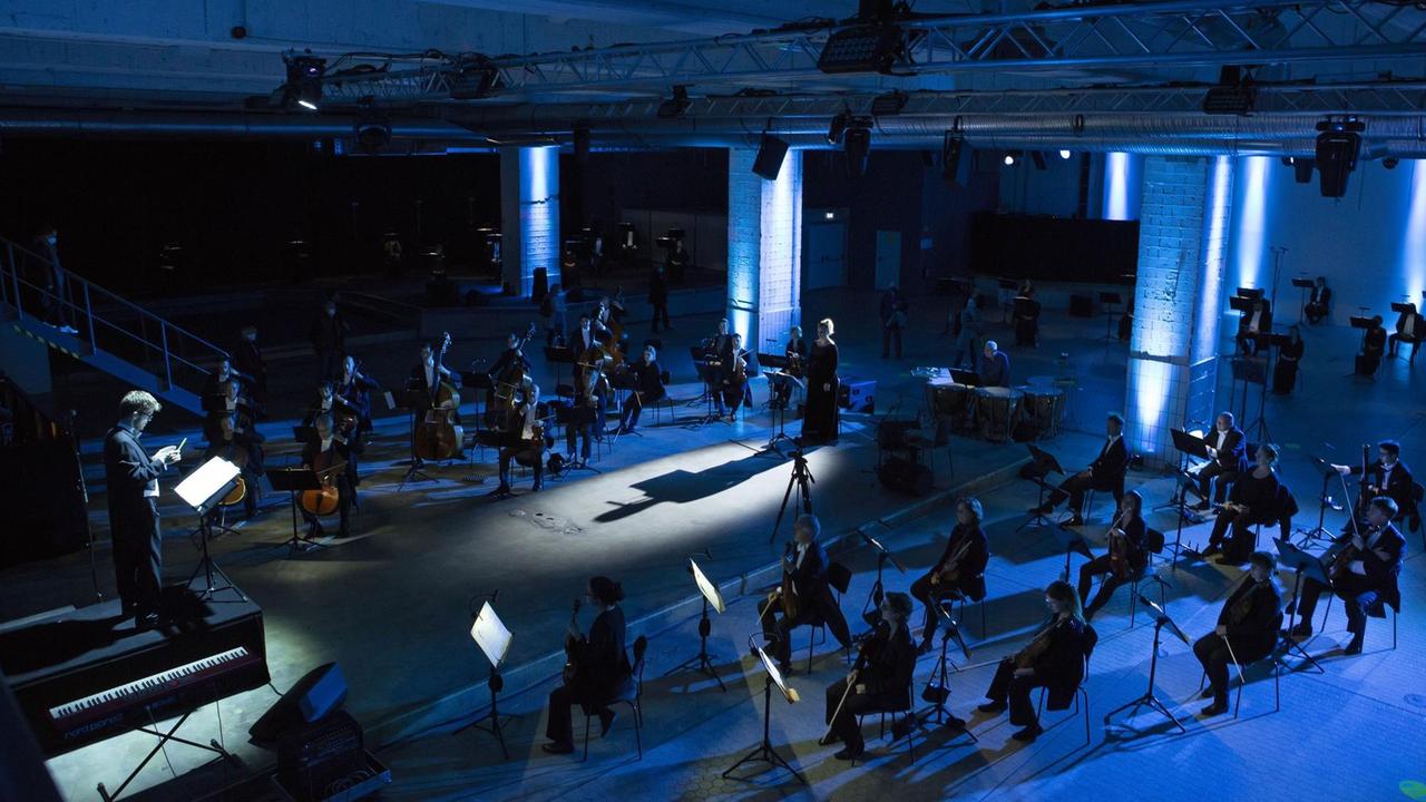 In einem blau ausgeleuchteten Industrieraum sitzt verstreut ein Orchester, das eine Sängerin weiträumig umringt samt Dirigenten, der auf einem erhöhten Pult steht. In einem blau ausgeleuchteten Industrieraum sitzt verstreut ein Orchester, das eine Sängerin weiträumig umringt samt Dirigenten, der auf einem erhöhten Pult steht.